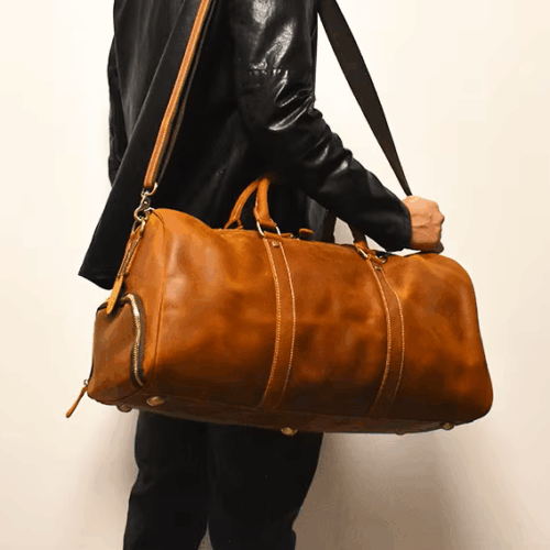 Ein Mann in schwarzer Lederjacke und Hose trägt vor einem schlichten, hellen Hintergrund einen großen braunen Weekender Herren Leder, Reisetasche und Sporttasche Herren mit Henkeln und Schultergurt.